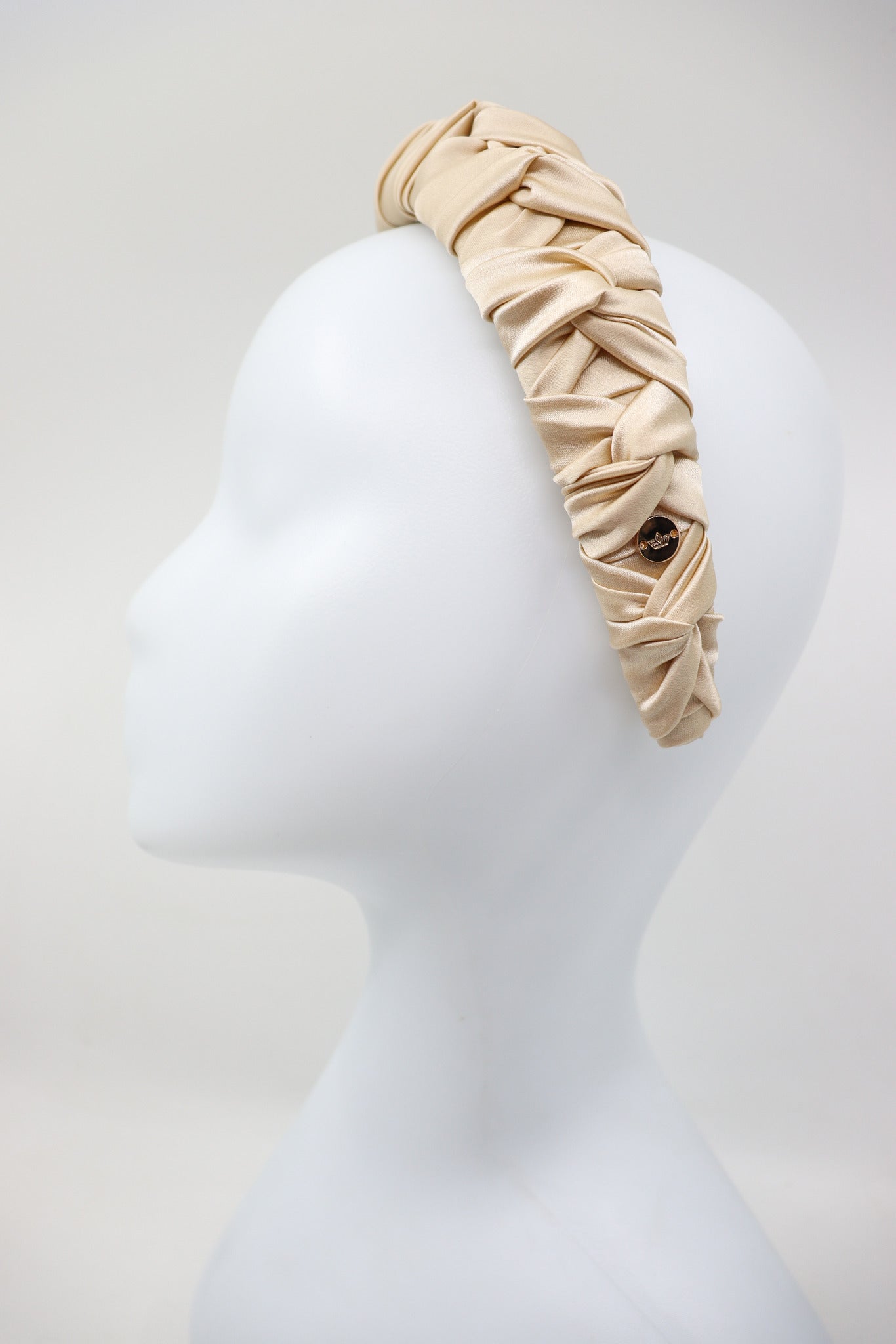 Close-up of the round metal logo tag on the left side of Jenna light beige silk headband.